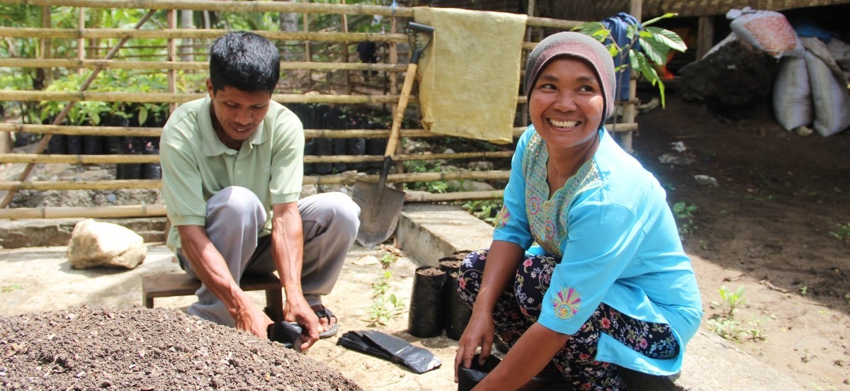 Woman in blue embroidered blouse smiles as she and a man next to her fill bags with dirt for planting